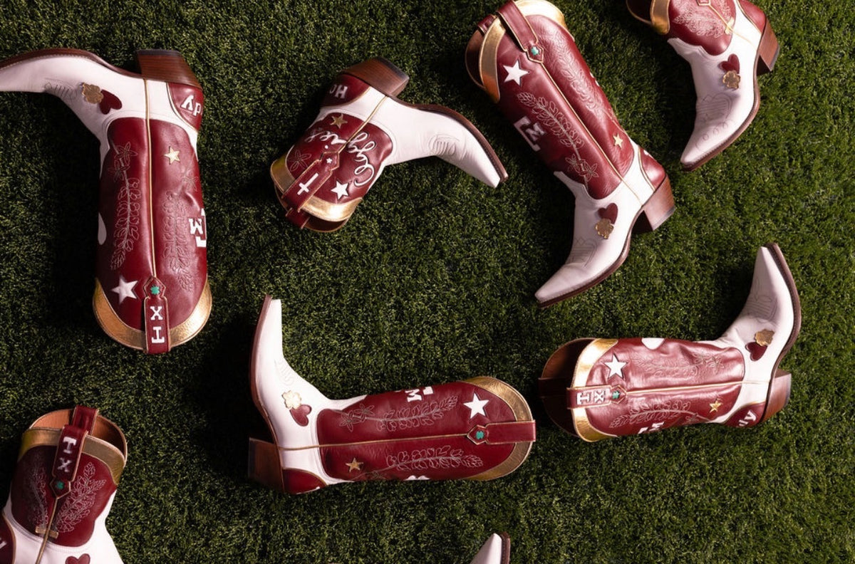 A group of Texas A&M western cowboy boots arranged on artificial green turf. The boots are primarily maroon and white with gold trim and embroidered star and heart details.