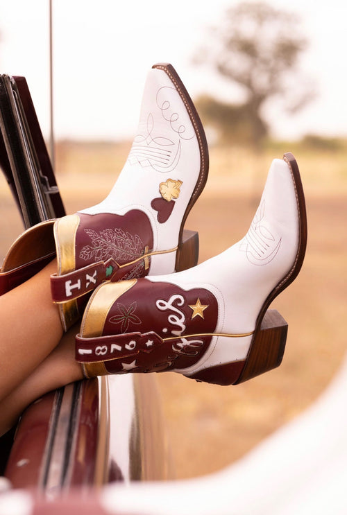 A pair of white cowboy boots with dark brown shafts and gold trim, featuring embroidered designs and the text 'Aggies' & '1876' on the straps, worn on a person's feet resting on a car door in an outdoor setting.